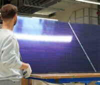 Ein Mitarbeiter hält in einer Fertigungshalle ein großformatiges bifaziales Photovoltaikmodul mit Perowskit-Silizium-Tandemzellen und Matrix-Schindel-Verschaltung.