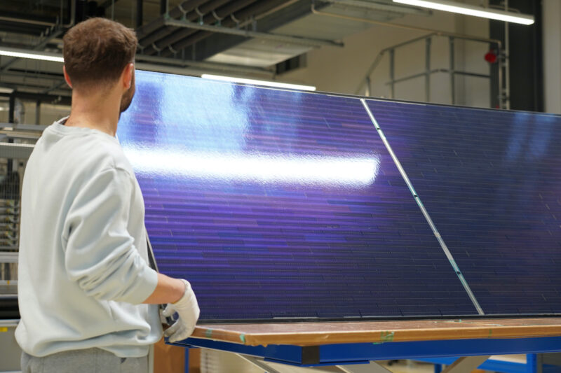 Ein Mitarbeiter hält in einer Fertigungshalle ein großformatiges bifaziales Photovoltaikmodul mit Perowskit-Silizium-Tandemzellen und Matrix-Schindel-Verschaltung.
