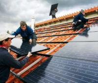 Mehrere Monteure installieren schwarze Photovoltaik-Module auf einem mit roten Ziegeln gedeckten Schrägdach.