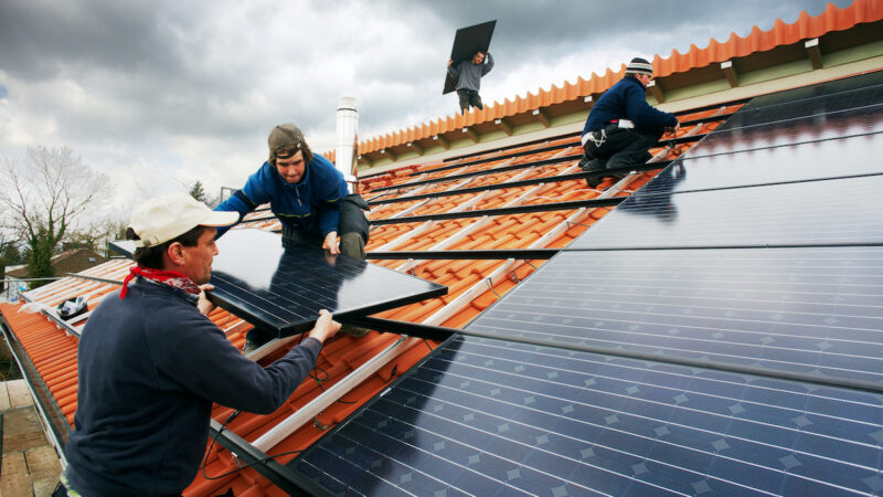 Mehrere Monteure installieren schwarze Photovoltaik-Module auf einem mit roten Ziegeln gedeckten Schrägdach.