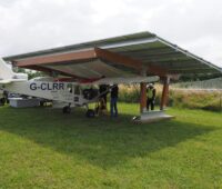 eine Art Photovoltaik-Carport auf einem grünen Rasen mit einem Leichtflugzeug darunter