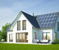 Weißes Einfamilienhaus, Neubau, mit Photovoltaik-Anlage und grünem Rasen vor blauem Himmel.