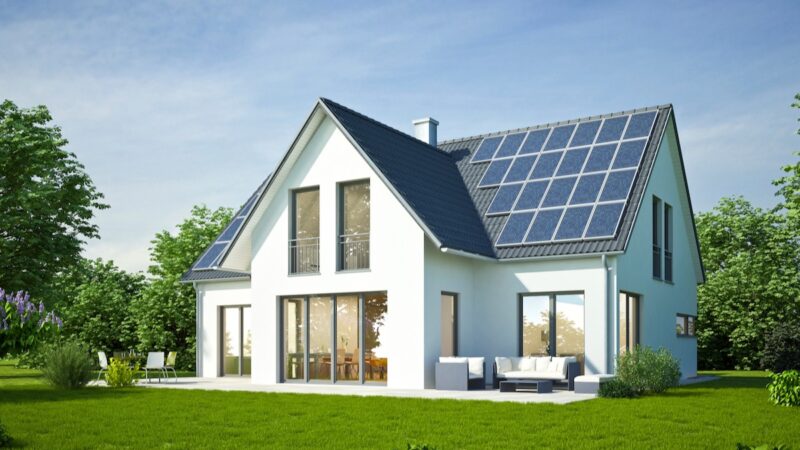 Weißes Einfamilienhaus, Neubau, mit Photovoltaik-Anlage und grünem Rasen vor blauem Himmel.