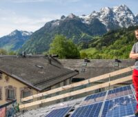 Photovoltaik-Anlagen in Österreich: PV-Module auf Dach, ein Installateur, im Hintergrund ein Kirchturm und Berge.