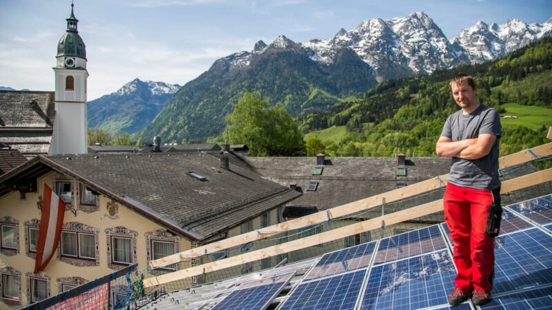 Photovoltaik-Anlagen in Österreich: PV-Module auf Dach, ein Installateur, im Hintergrund ein Kirchturm und Berge.