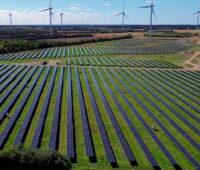 Luftbild einer Photovoltaik-Freiflächen-Anlage, im Hintergrund Windräder