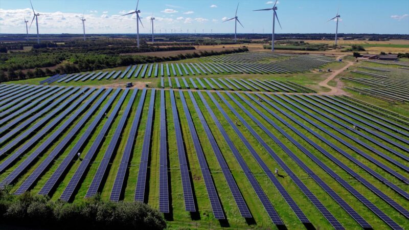 Luftbild einer Photovoltaik-Freiflächen-Anlage, im Hintergrund Windräder