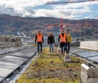 Photovoltaik-Anlage auf grünem Dach, zwischen den Modulreihen Menschen, im Hingergrund Hügel mit Wald