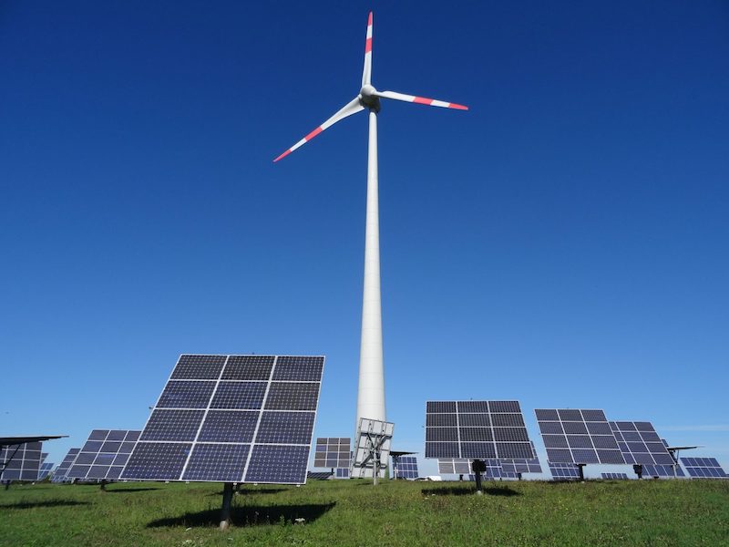 Eine Windenergie-Anlage und mehrere Photovoltaik-Nachführsysteme auf einer Wiese vor blauem Himmel