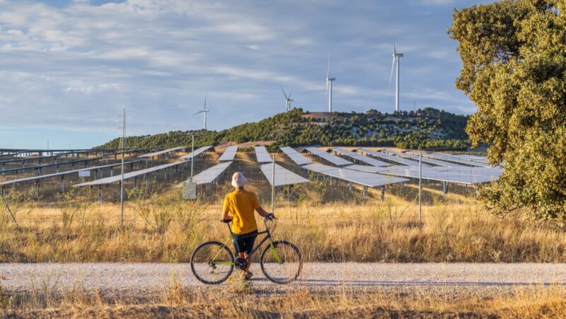 Mensch mit Fahhrad steht auf einem Weg und schaut zu Solarpark und Windenergie-Anlagen. Symbolbild für Photovoltaik und Windenergie in der Region Kastilien und Leon, Spanien.