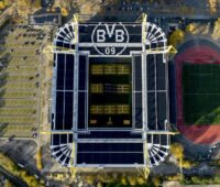Senkrechte Luftaufnahme eines Fußballstadions. Auf den Dachflächen sind großflächig Photovoltaikmodule installiert. Neben dem Stadion sind Parkflächen und eine Leichtathletikanlage zu sehen.