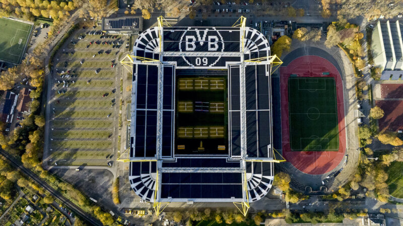 Senkrechte Luftaufnahme eines Fußballstadions. Auf den Dachflächen sind großflächig Photovoltaikmodule installiert. Neben dem Stadion sind Parkflächen und eine Leichtathletikanlage zu sehen.