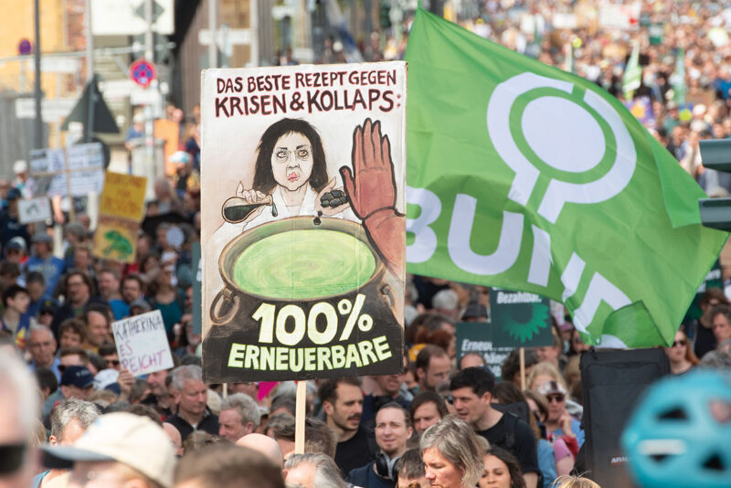 Demonstrationszug für Energiewende in Berlin - Vorderung ein gemaltes Plakat das Katherina Reiche als Giftmischerin zeigt, die den grünen Stromsee mit Öl und Kohle vergiftet.