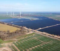 Luftbild eines PV-Parks auf grüner Wiese und Windenergie im Hintergrund.