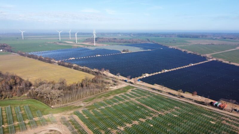 Luftbild eines PV-Parks auf grüner Wiese und Windenergie im Hintergrund.
