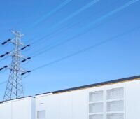 Ein im Vordergrund angeschnittener Batteriespeicher PowerTitan 2.0 in lichtgrau vor blauem Himmel. Im Hintergrund eine Strommast.