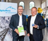 Die beiden Verantwortlichen stehen nach der Vertragsunterzeichnung nebeneinander vor einem Werbebanner von AllgäuNetz. Einer zeigt einen digitalen Zähler.