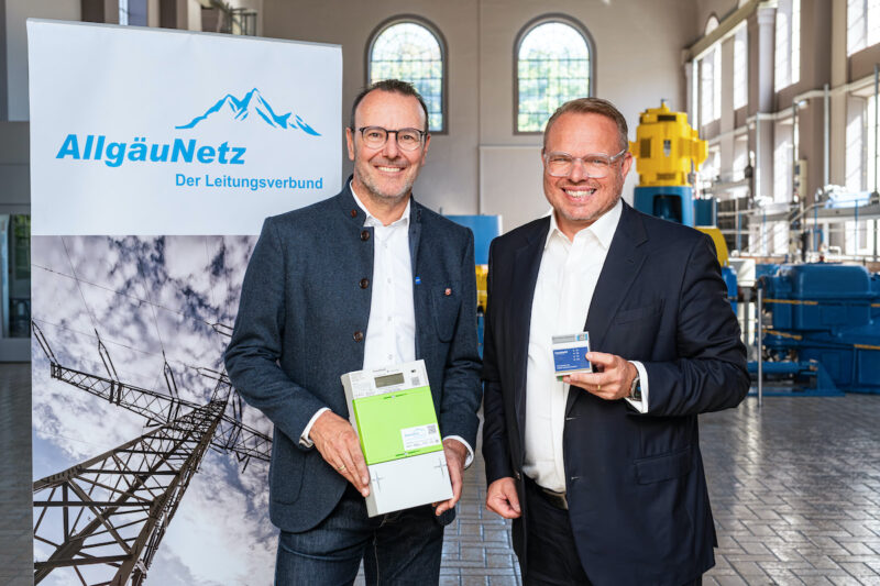 Die beiden Verantwortlichen stehen nach der Vertragsunterzeichnung nebeneinander vor einem Werbebanner von AllgäuNetz. Einer zeigt einen digitalen Zähler.