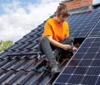 Installateurin montiert sitzend auf einem Schrägdach Solarmodule.