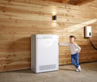 Ein Kind lehnt mit einer Hand an einer hölzernen Carport-Wand neben einem Energiespeicher.