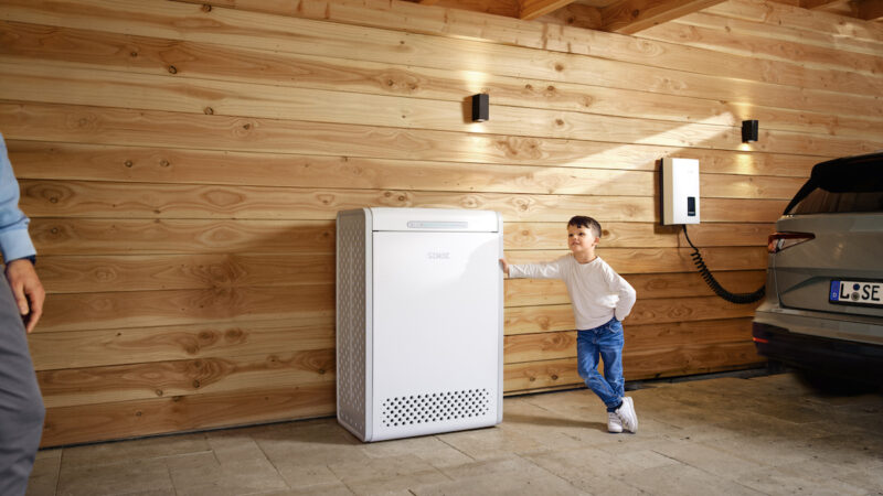 Ein Kind lehnt mit einer Hand an einer hölzernen Carport-Wand neben einem Energiespeicher.