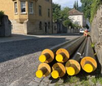Fernwärmeleitungen liegen am Rande einer Straße bereit. Im Hintergrund ältere Gebäude
