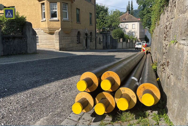 Fernwärmeleitungen liegen am Rande einer Straße bereit. Im Hintergrund ältere Gebäude