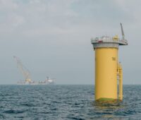 Fundament für Offshore-Windenergieanlage, das aus dem Wasser ragt - im Hintergrund Transportschiff.