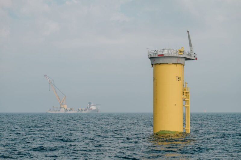 Fundament für Offshore-Windenergieanlage, das aus dem Wasser ragt - im Hintergrund Transportschiff.