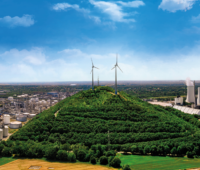 Blick in die Landschaft aus Vogelperspektive: links Stadt, Mitte Wald mit Windrädern, rechts Raffinerie vor blauem Himmel.
