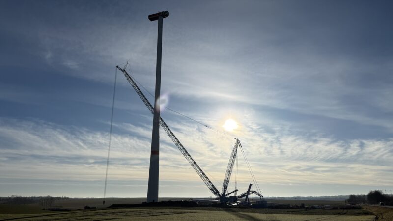 Vor einer milchigen Sonne finden Arbeiten mit einem Kran an einer Windenergieanlage statt.