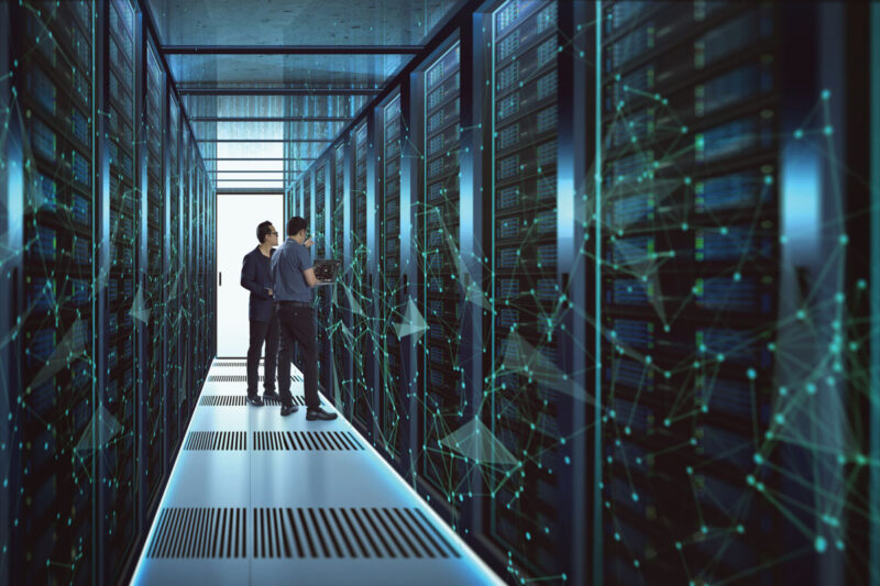 Zwei Männer im Gang eines Rechenzentrums zwischen Computer- oder Speicher-Racks hinter Glas, wobei das Foto im bläulichen Licht an eine Science-Fiction-Umgebung erinnert.