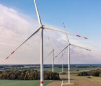 Mehrere Windenergieanlagen mit rot markierten Rotorblättern stehen zwischen Feldern und Waldflächen.