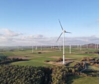 Luftaufnahme eines Windparks in einer landwirtschaftlich geprägten Hügellandschaft. Mehrere Windenergieanlagen stehen zwischen Feldern und Waldflächen. Im Vordergrund ist eine moderne Windkraftanlage zu sehen, im Hintergrund weitere Anlagen.