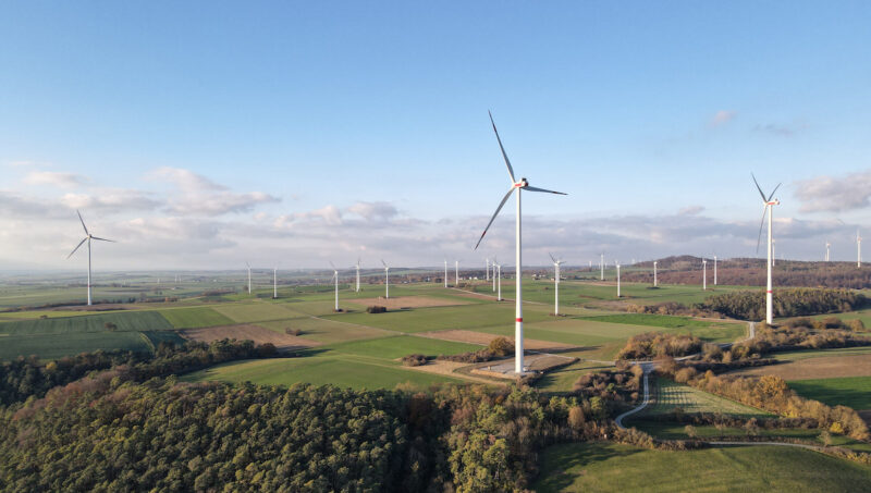 Luftaufnahme eines Windparks in einer landwirtschaftlich geprägten Hügellandschaft. Mehrere Windenergieanlagen stehen zwischen Feldern und Waldflächen. Im Vordergrund ist eine moderne Windkraftanlage zu sehen, im Hintergrund weitere Anlagen.