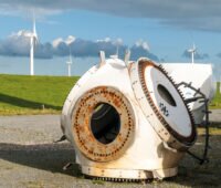 Windkraft-Repowering-Projekt in Nordfriesland. Im Hintergrund verschiedene Generationen von Windenergieanlagen und ein Nordseedeich. Im vordergrund die leicht angerostete Nabe einer demontierten Windenergieanlage.