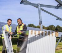 Zwei Männer mit einem Paket Solarmodule neben einem Freiflächen-Montagegestell im Solarpark - Symbolbild für Revamping