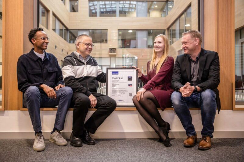 Vier Personen in einem Bürogebäude sitzend mit Zertifikatsurkunde.