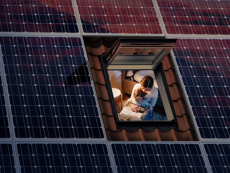 Offenes Dachfenster von oben eingerahmt mit Photovoltaikmodulen mit Blick auf Frau und Kind.