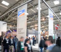 Das Foto zeigt einen Messestand in einer Halle mit davor vorbeilaufenden Menschen, die durch die Bewegung verschwommen sind.