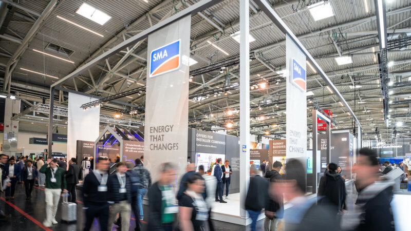 Das Foto zeigt einen Messestand in einer Halle mit davor vorbeilaufenden Menschen, die durch die Bewegung verschwommen sind.