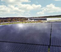 Eine Freiflächen-Solaranlage, auf der die Sonne reflektiert, vor einem Werksgelände.