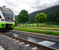 Eine Zugstrecke: im Vordergrund die Schienen, zwischen denen Photovoltaikmodule installiert sind, im Hintergrund ein Zug, der über die Schienen fährt.