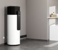 Weiße, zylindrische Brauchwasser-Wärmepumpe WWK-I 300 Plus in einem Hauswirtschaftsraum neben Waschmaschine und Trockner. Links ist das Stiftung-Warentest-Siegel „Sehr gut (1,5)“ eingeblendet.