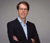Portraitfoto von Holger Schneidewindt mit verschränkten Armen im dunklen Blazer vor grauem Hintergrund