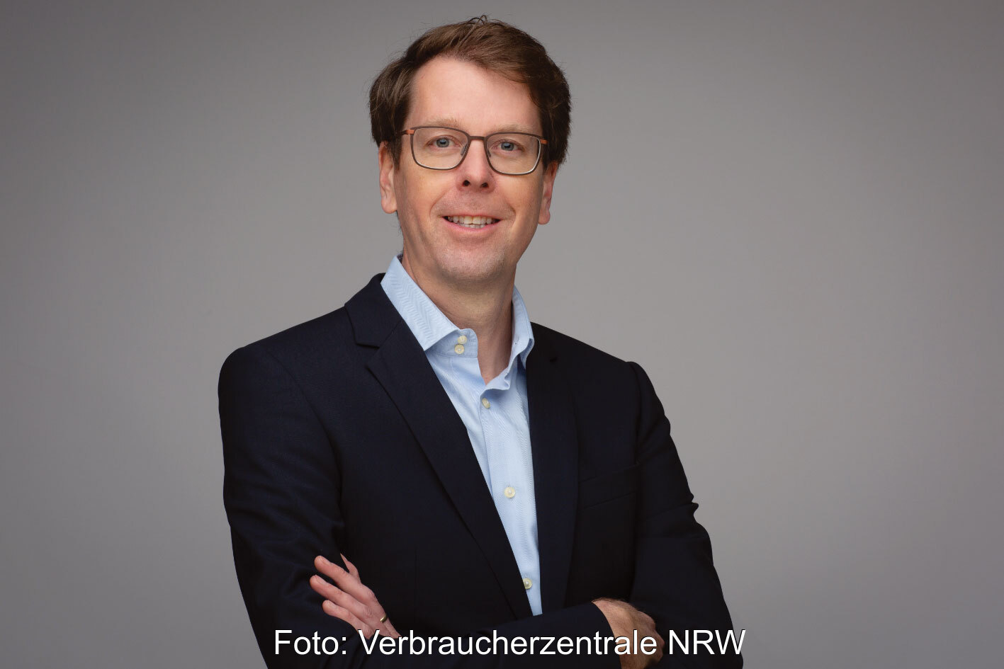 Portraitfoto von Holger Schneidewindt mit verschränkten Armen im dunklen Blazer vor grauem Hintergrund
