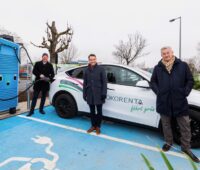 Vier Männer in schwarzen Jacken stehen neben der blauen Schnelladesäule vor einem weißen E-Auto.