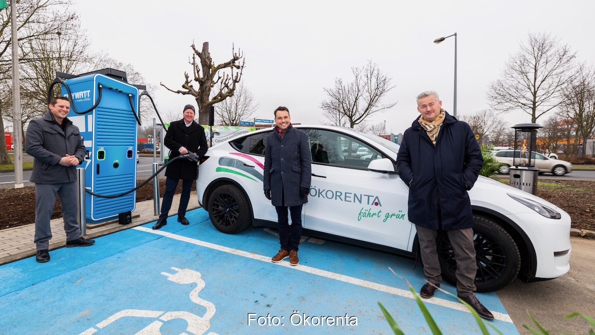 Vier Männer in schwarzen Jacken stehen neben der blauen Schnelladesäule vor einem weißen E-Auto.