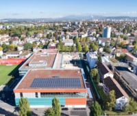 Luftaufnahme des Schweizer Ortes Allschwil. Im Vordergrund eine Schulturnhalle mit einer PV-Anlage auf dem Flachdach.