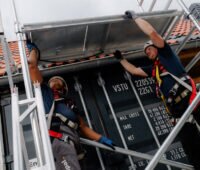 Zwei Monteure mit Schutzhelmen, Handschuhen und Auffanggurten heben eine Metallplattform innerhalb eines Gerüstaufbaus an einem ziegelgedeckten Steildach an.